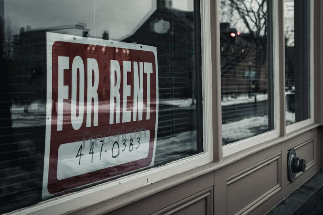 A red "for rent" sign in a window of a grey building